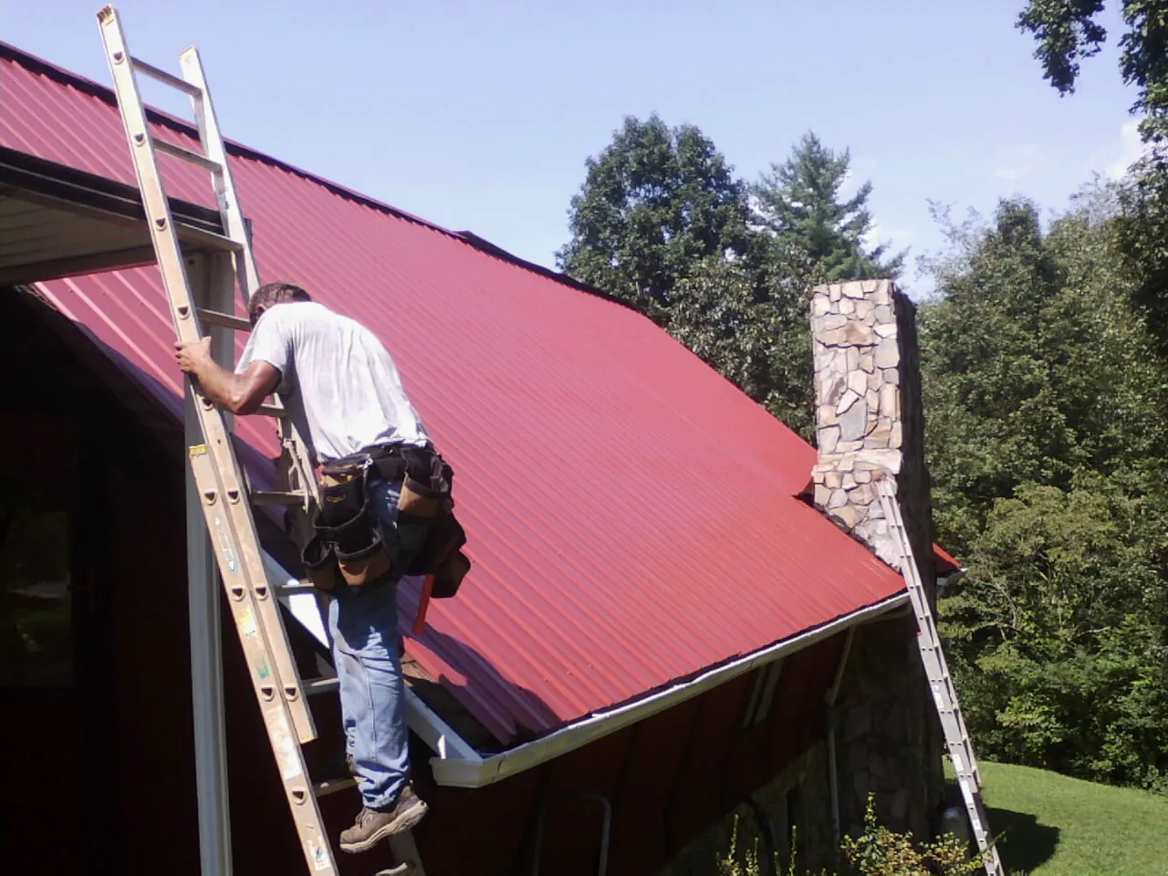 Roofer on a ladder beside a red metal roof and stone chimney — Soffit & Fascia Repair by Roofing Brothers in Maine
